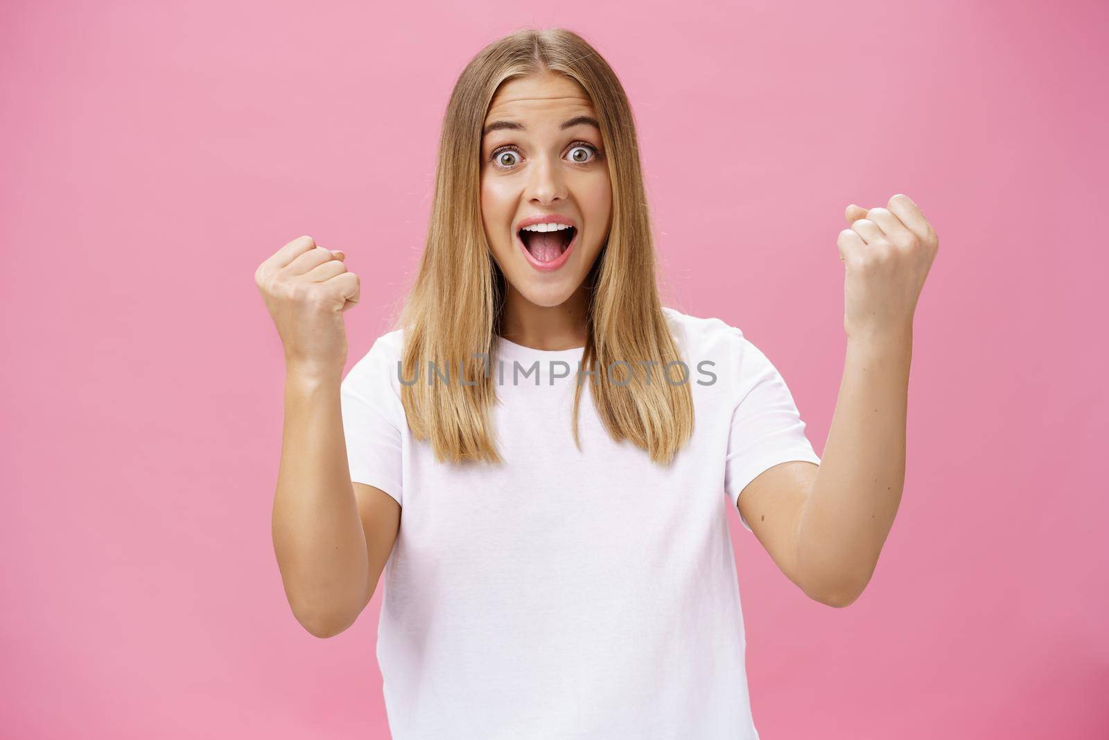Excited cheerful and optimistic charming woman with fair hair in white t-shirt raising fists in victory and triumph yelling yes in success and amazement standing supportive against pink background. Lifestyle.