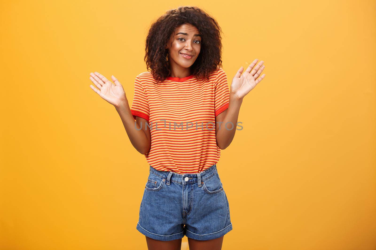 No idea do not care. Careless and indifferent calm happy african american woman with curly hair in summer clothes raising palms and shrugging in uninvolved pose smiling carefree being unaware. Body language concept