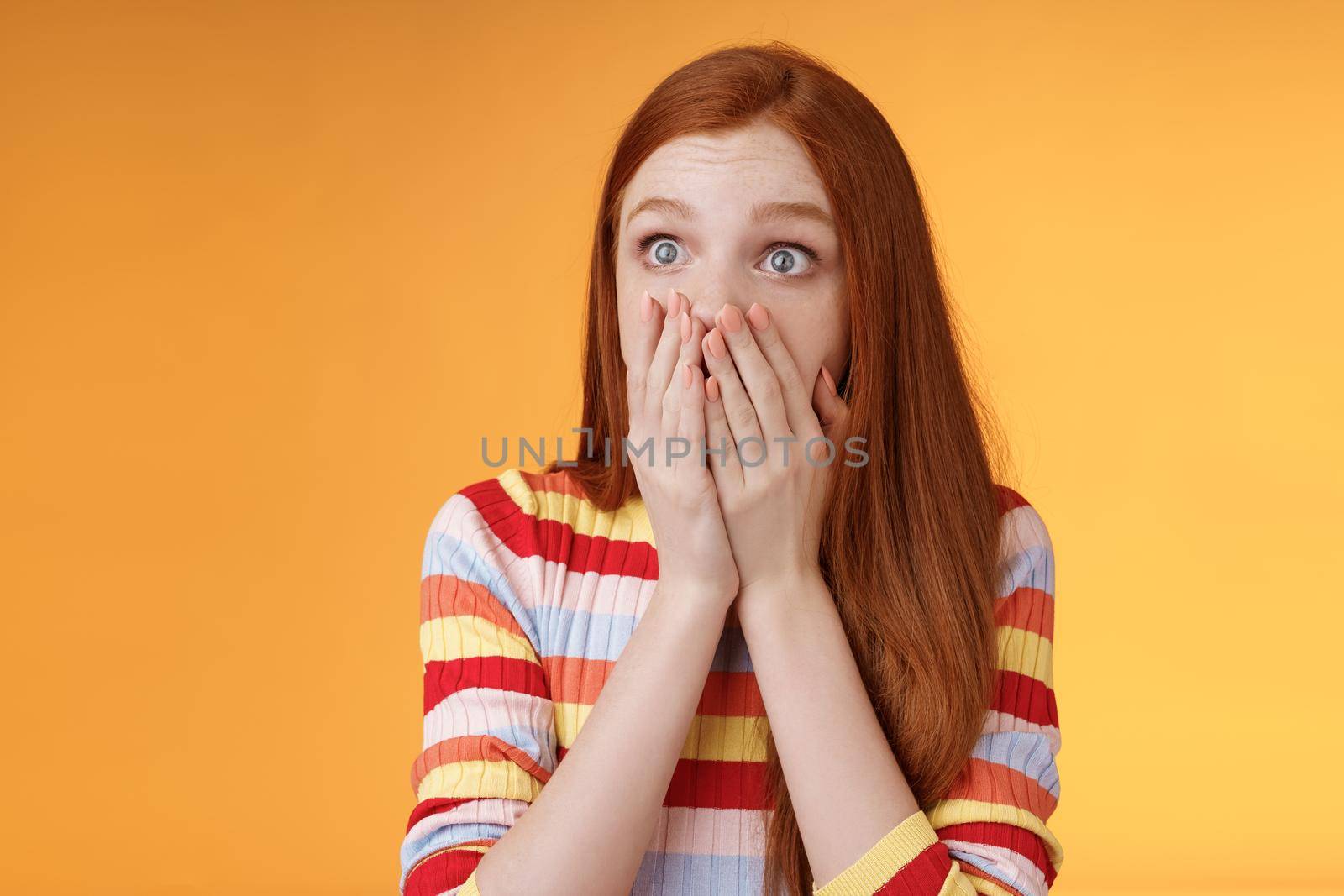 Shocked upset redhead female witness terrible accident gasping cover mouth palms astonished staring left frightened express empathy worry standing orange background panicking. Copy space