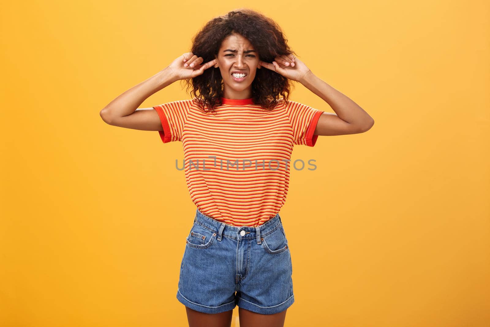 What that anoying sound. Disturbed and displeased dark-skinned girl with curly hair frowning clenching teeth from discomfort closing ears with index fingers from loud irritating sound over orange wall by Benzoix