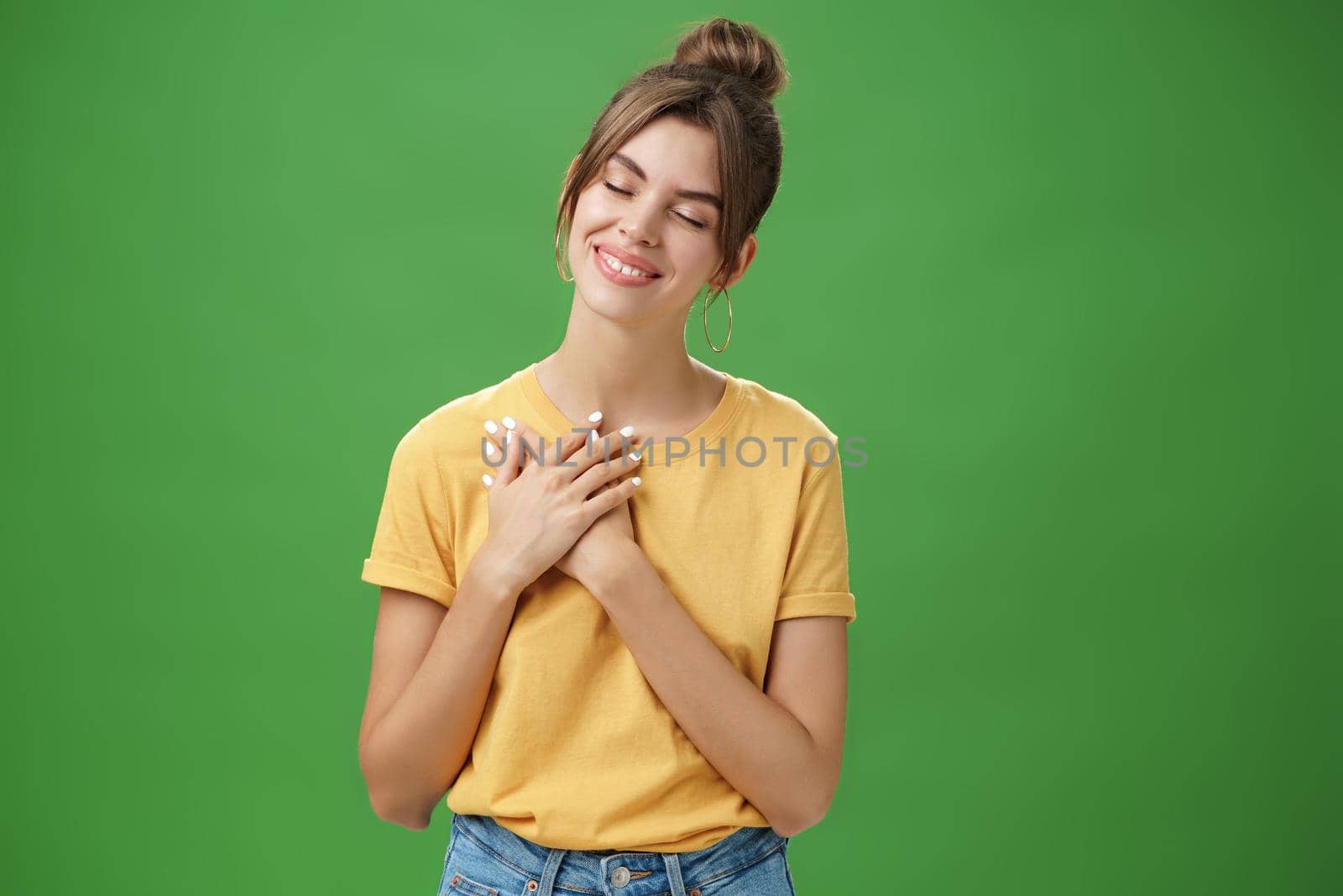 Happy kind and timid young attractive woman with gapped teeth smiling sincere and grateful closing eyes pressing hands on chest in thankful pose, daydreaming, recalling nice tender memories.