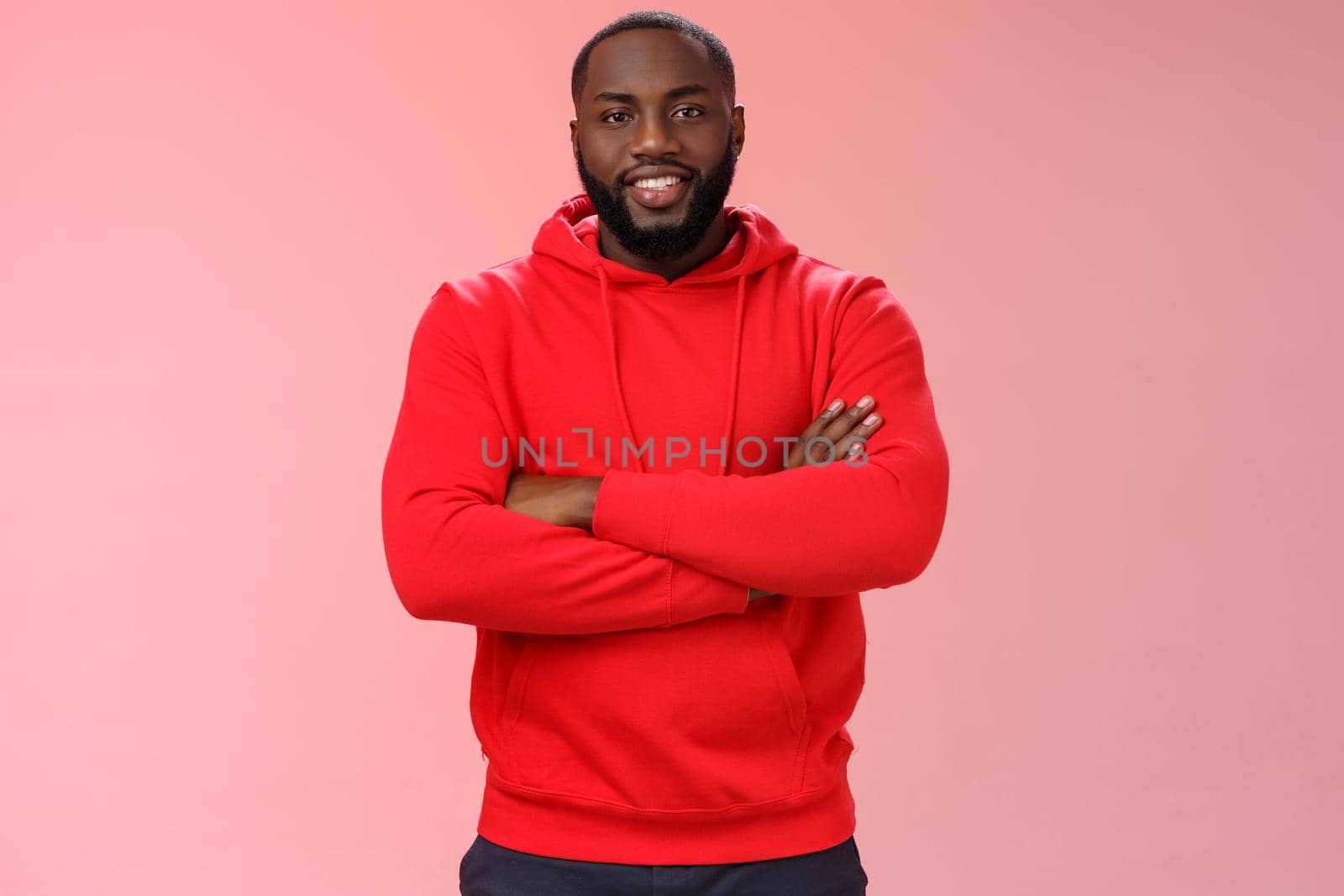 Attractive bearded african-american young guy wearing red hoodie cross arms chest smiling friendly showing friends new place grinning delighted buying own flat, standing proud pink background.