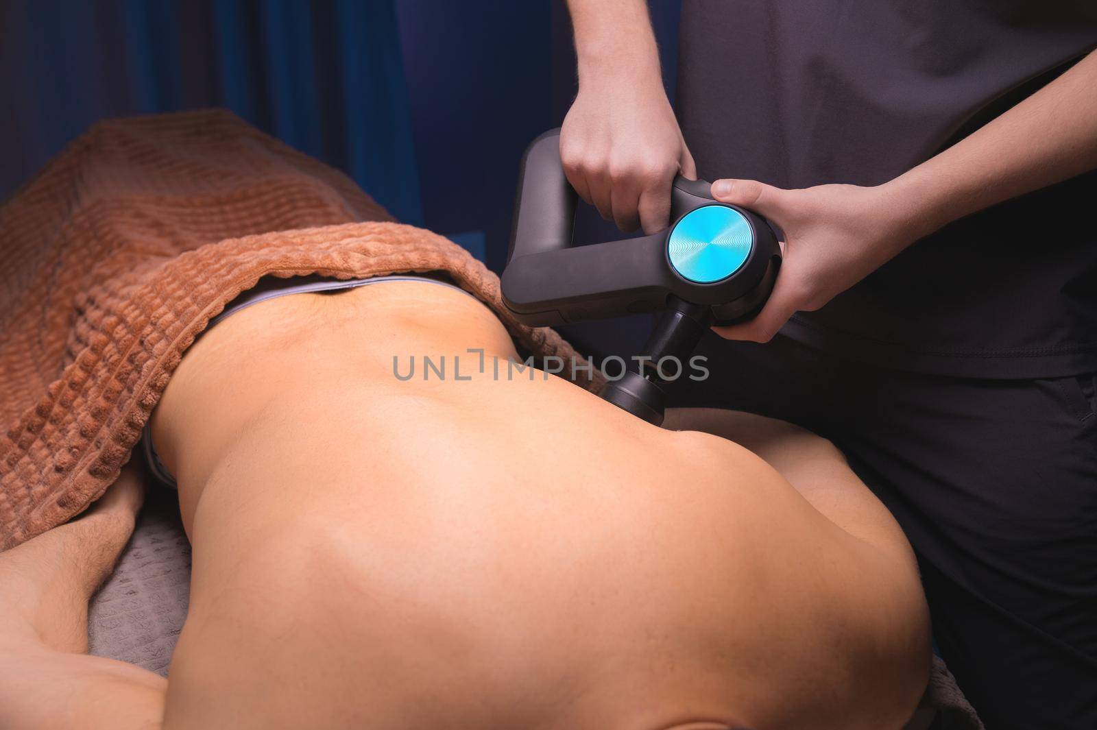 Close-up of a professional male masseur stimulates the back muscles of a male patient in a dark spa room for massage. Percussion mechanical effect on overstrained muscles by yanik88