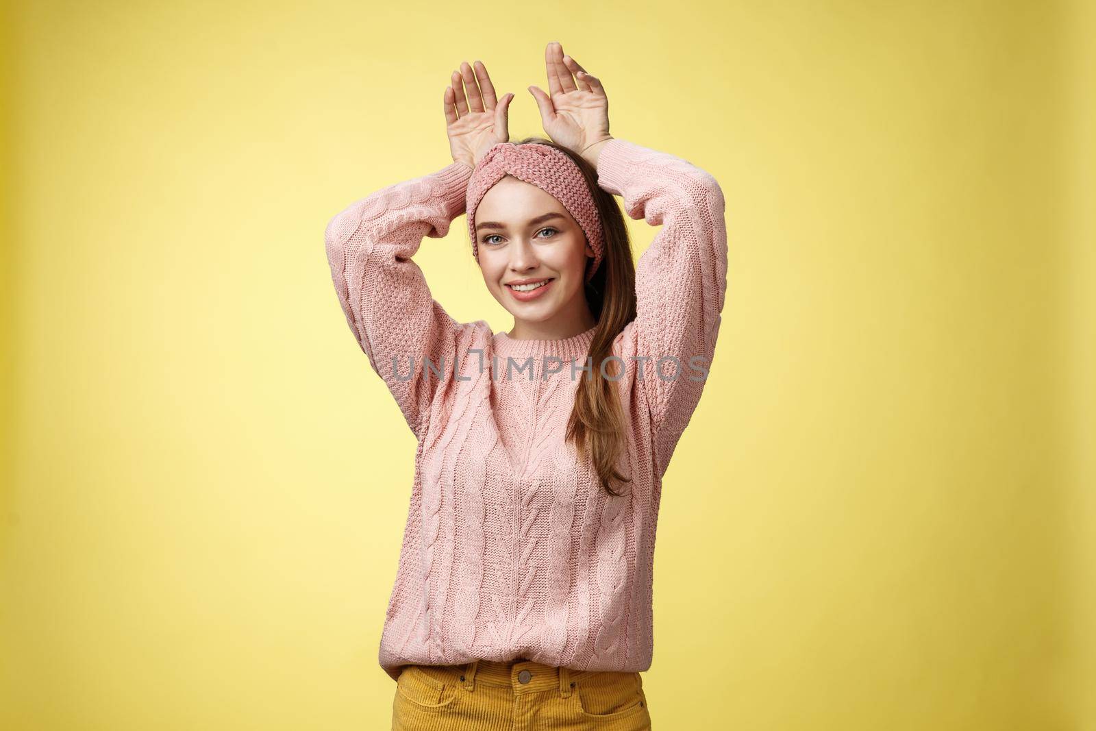 Nice charming playful young european female having fun, playing mimicking bunny showing rabbit ears with hands behind head, smiling cute, tender, happy greeting boyfriend welcome back ove yellow wall.