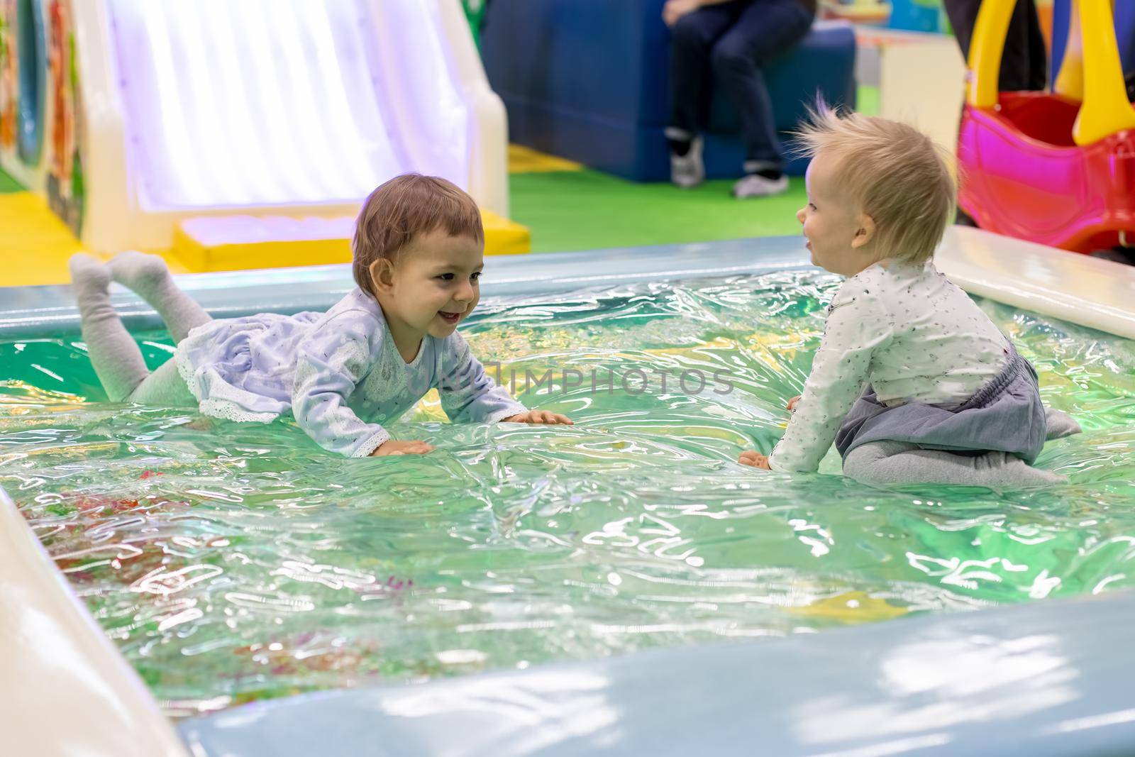 two little cute baby toddler girls in a dress sit, lie and laugh enjoying in a toy pool on a children's playground of day care play room, soft focus, background in blur