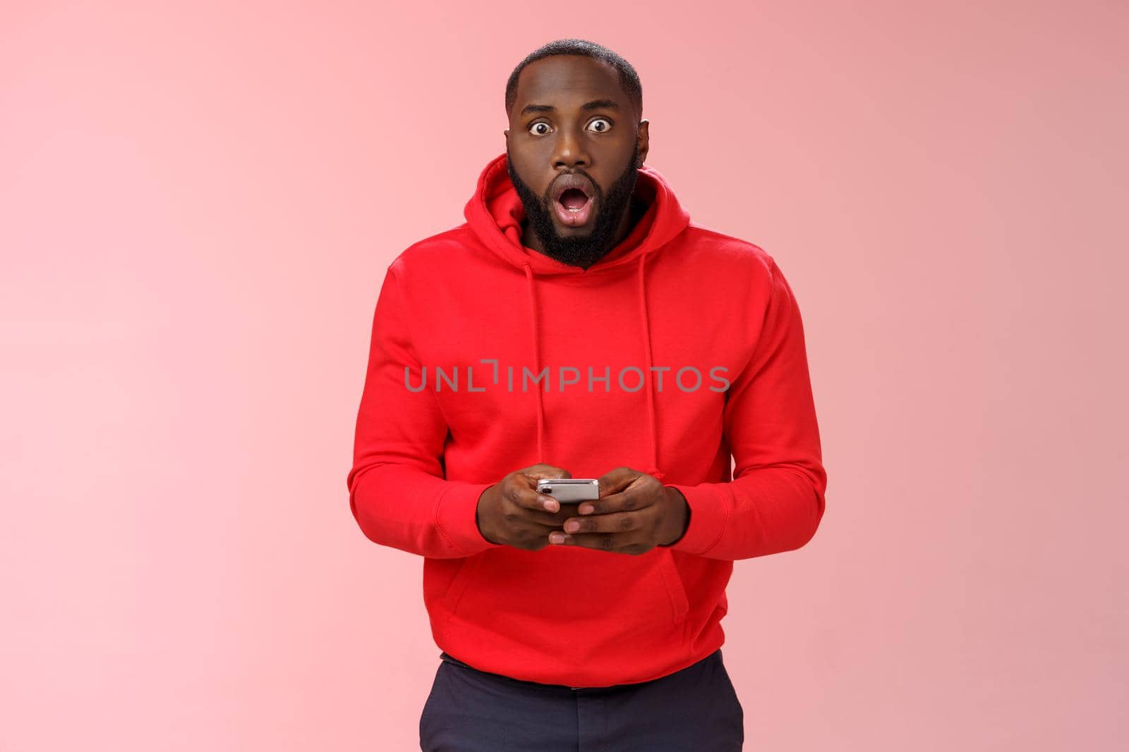 Amused overhwelmed black bearded guy in red hoodie drop jaw gasping widen eyes staring camera impressed holding smartphone using amazing cool game astonished new phone features, standing pink wall by Benzoix