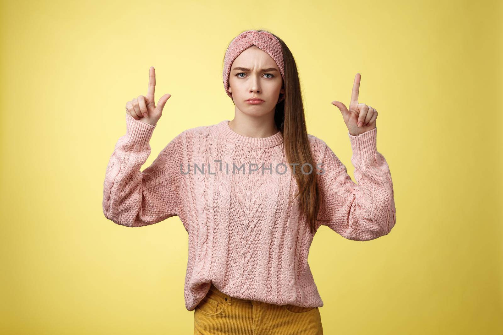 Moody disappointed and upset unamused cute young european girl in knitted warm sweater headband expressing dislike and disagreement pointing up, sulking frowning displeased unhappy with bad choice.