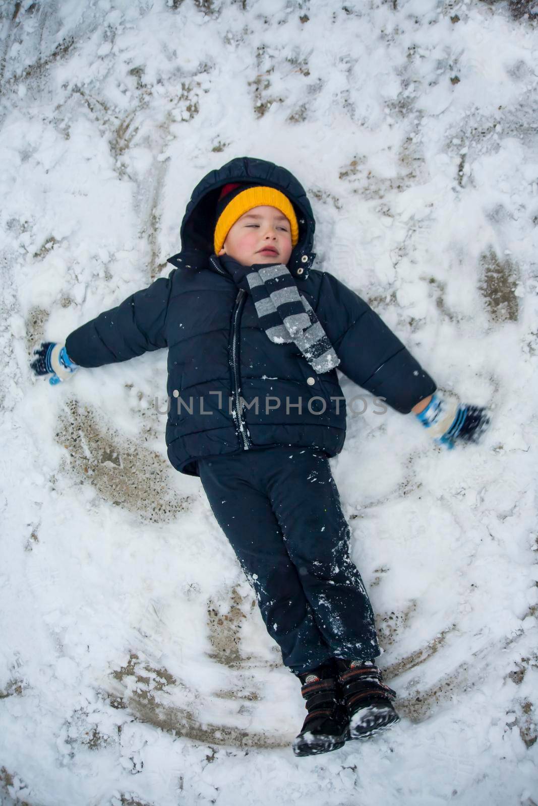 The boy in a blue jacket and blue pants lies in the snow and makes an angel. His yellow hat comes to the fore. The play and joy of a child in the snow.