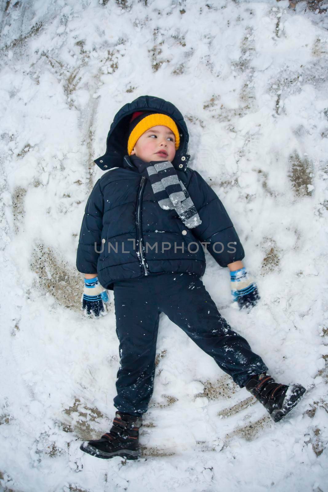 The boy in a blue jacket and blue pants lies in the snow and makes an angel. His yellow hat comes to the fore. The play and joy of a child in the snow.