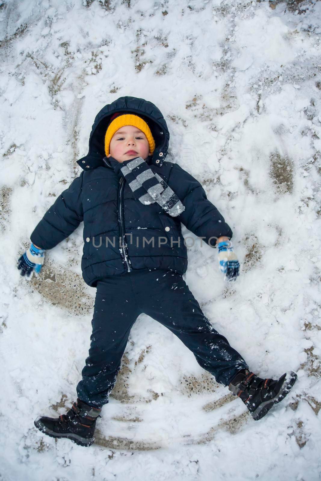 The boy in a blue jacket and blue pants lies in the snow and makes an angel. His yellow hat comes to the fore. The play and joy of a child in the snow.