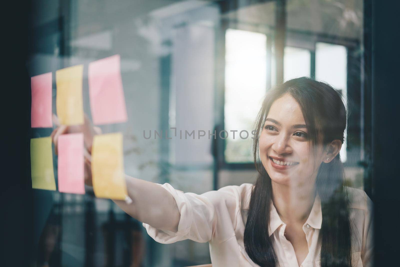 Young creative woman professional using sticky notes in glass wall to writing strategy business plan to development grow to success.