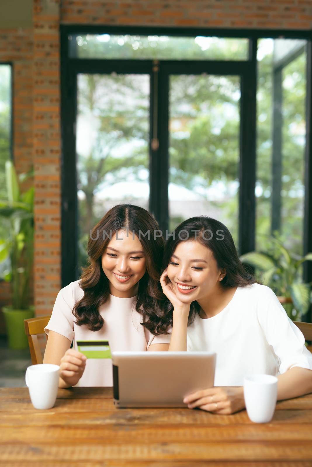 Happy relaxed young female friends doing online shopping through laptop and credit card in living room by makidotvn
