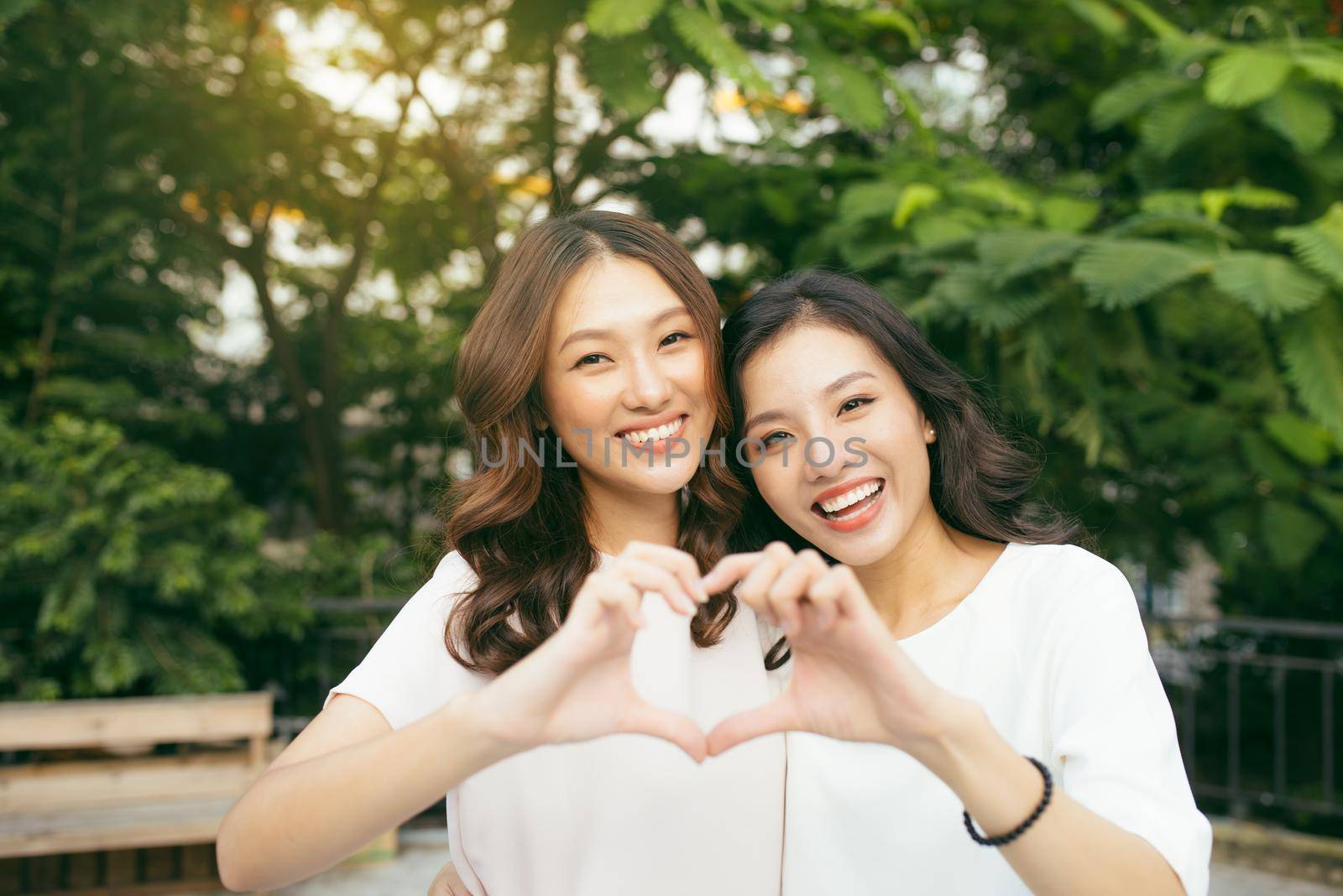 Two girl friends standing in a hug and forming heart shape with hands in the nature. Friendship concept