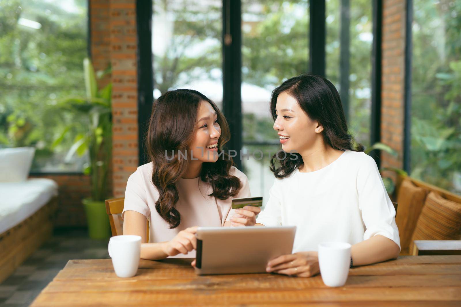 Happy relaxed young female friends doing online shopping through tablet and credit card in living room