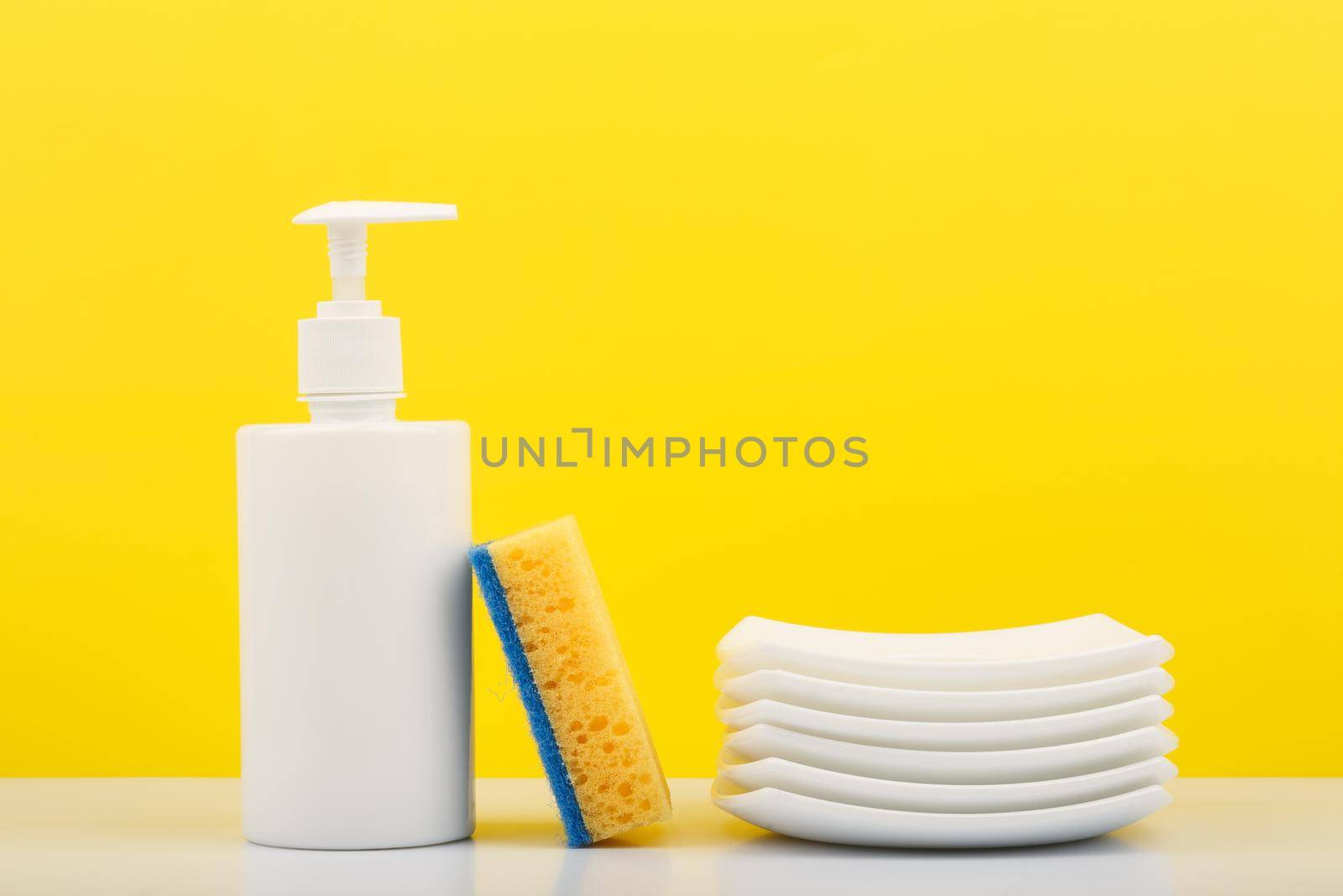 Colorful dishwashing concept, creative composition with liquid detergent in white plastic bottle with dispenser, yellow cleaning sponge and pile of clean plates against yellow background