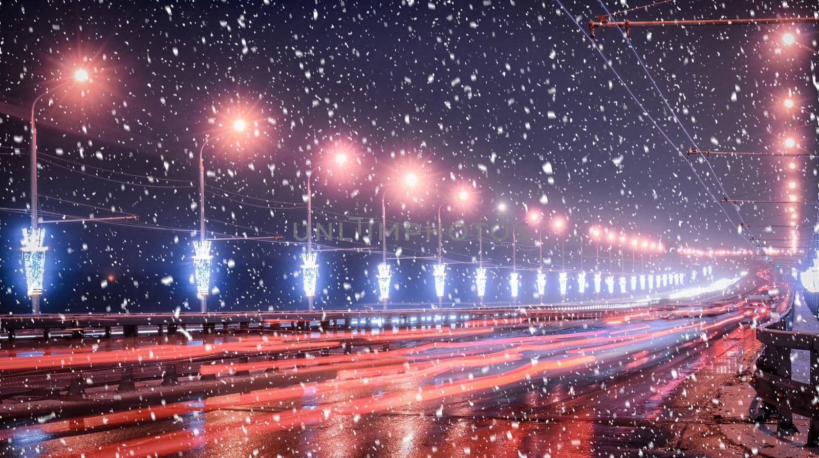 Traces of headlights from cars moving at winter night on the bridge, illuminated by lanterns in a snowfall. Lights reflecting in the wet asphalt.