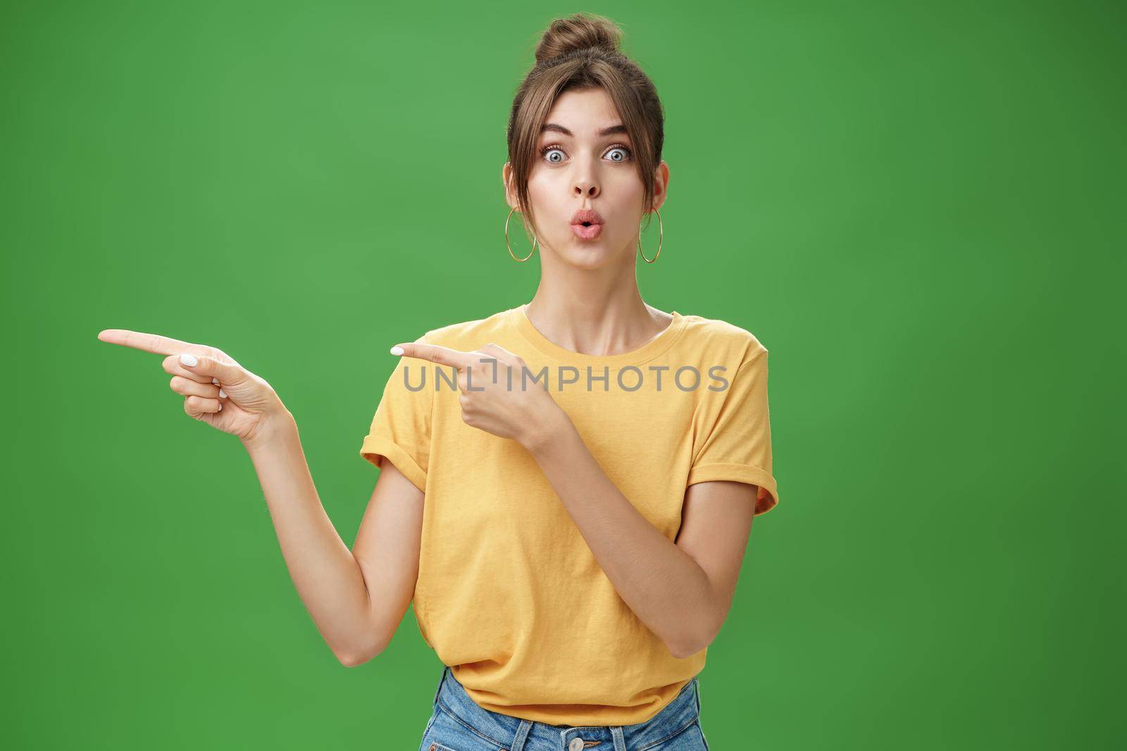 Woman reacting to awesome sales in near shop pointing left and gazing impressed and excited at camera folding lips from desire and amazement staring at camera thrilled posing over green background. Lifestyle.