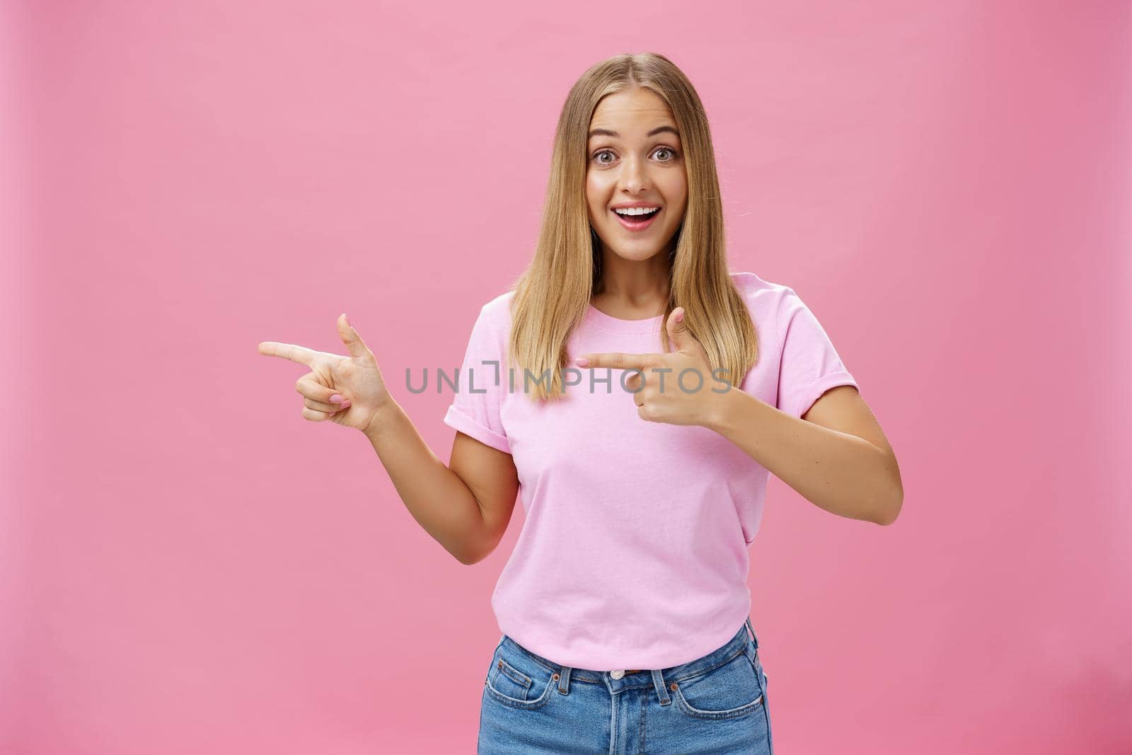 Portrait of charismatic and enthusiastic good-looking female with blonde hair and tanned skin in casual t-shirt pointing left smiling amused and delighted standing impressed over pink background. Lifestyle.