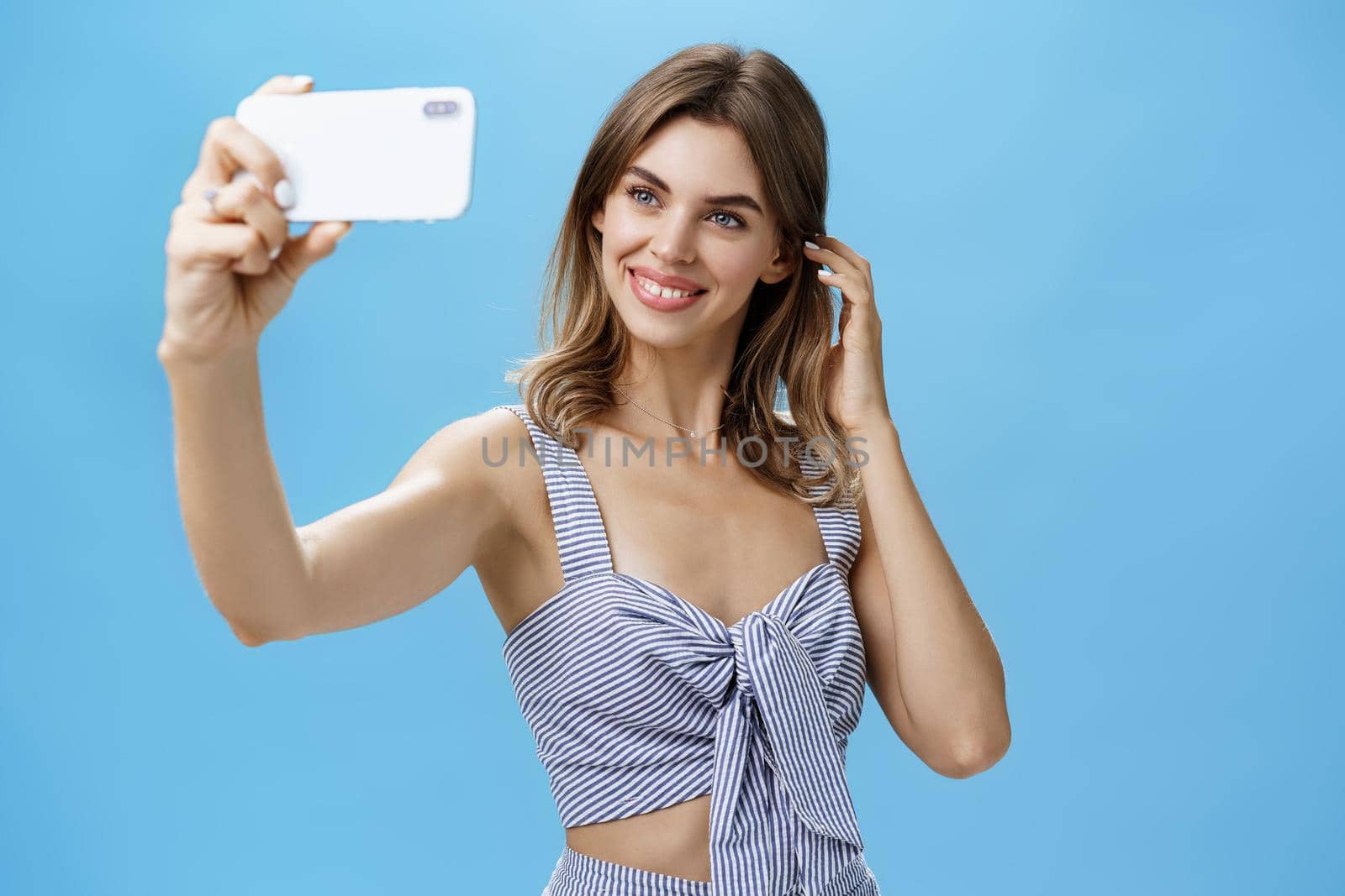 Attractive woman with good self-esteem in stylish matching outfit flicking hair behind ear smiling joyfully at smartphone screen, taking selfie to post in internet and gain followers over blue wall. Lifestyle concept