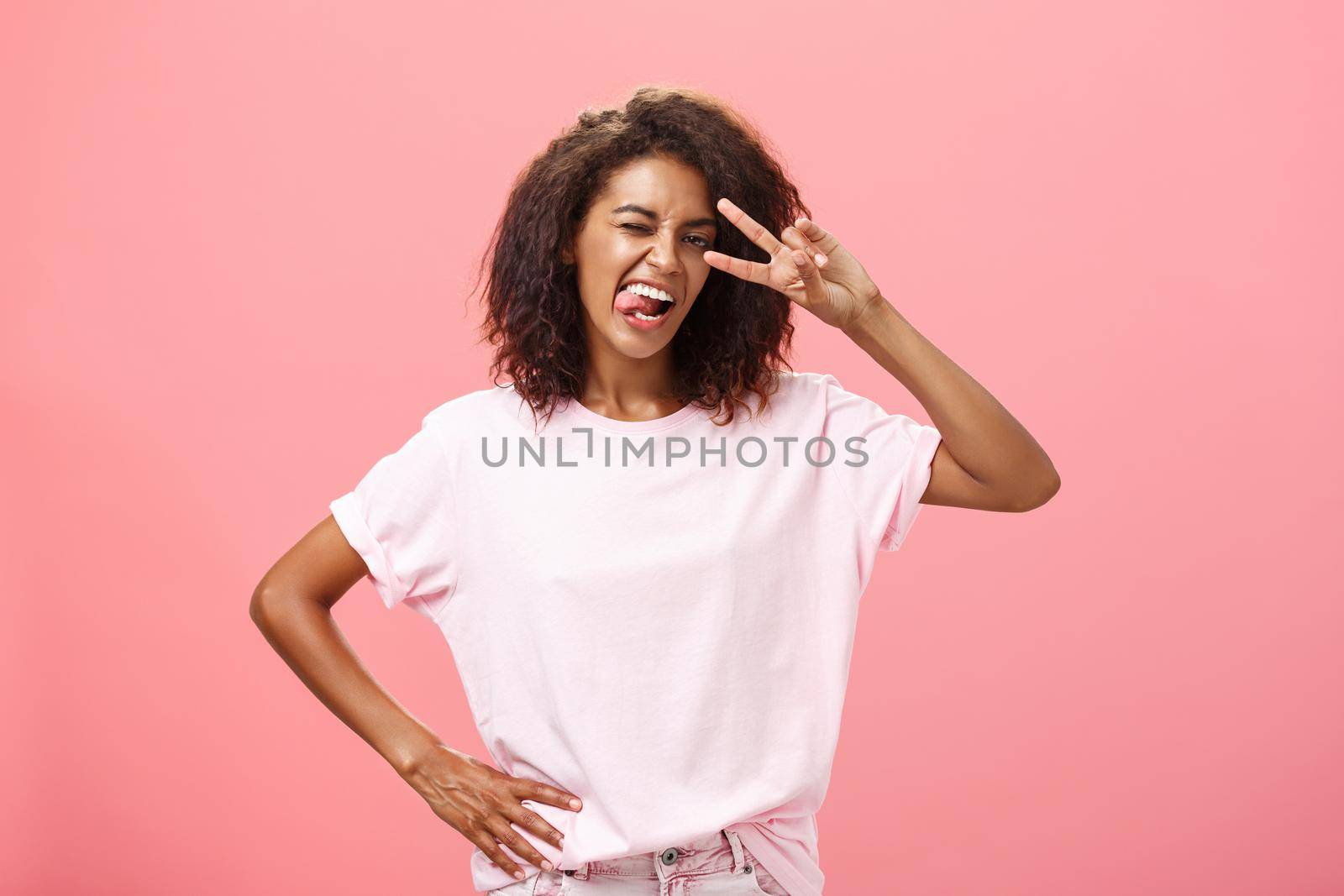 Not afraid express myself. Joyful charismatic african american woman in t-shirt with afro haircut showing tongue playfully and daring making peace sign over eye and winking posing over pink background.
