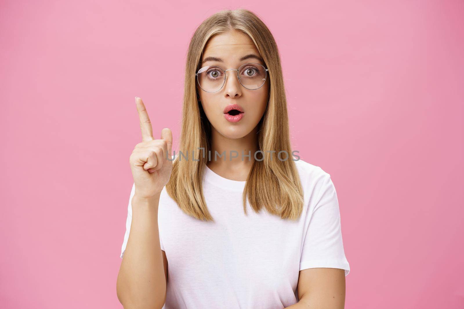 One more thing need said. Intelligent and smart female A student in glasses and white t-shirt open mouth and folding lips raising index finger in eureka gesture adding suggestion, having idea. Body language concept