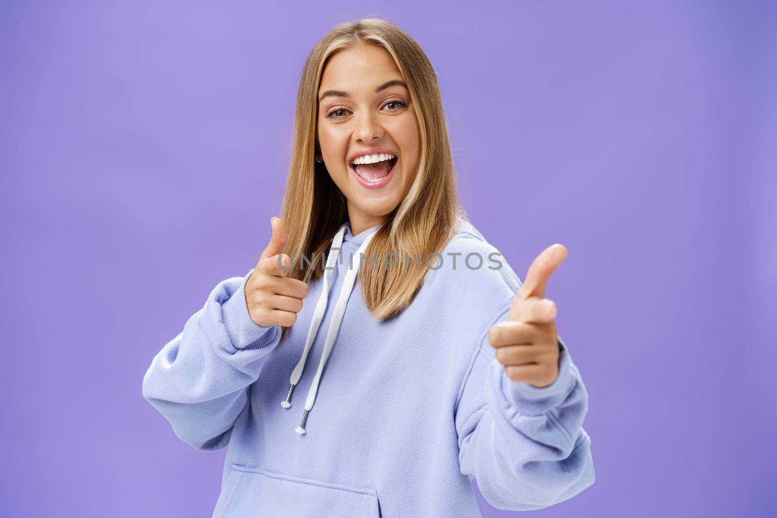 Enthusiastic and charismatic woman feeling joyful meeting friends checking out cool copy space pointing with finger guns at camera smiling broadly posing in stylish hoodie over purple wall. Copy space