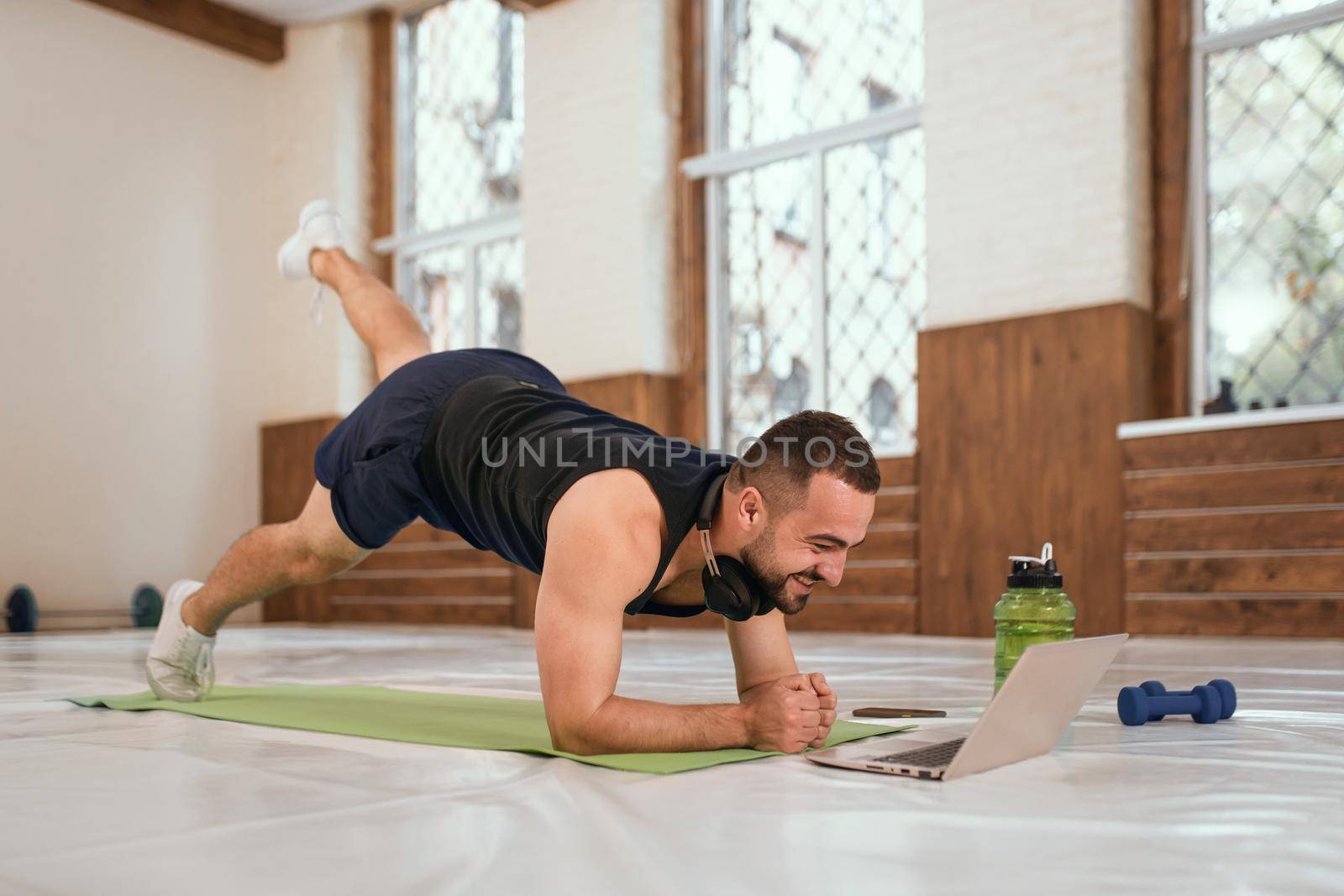Handsome young sport man doing one leg up push ups exercise in empty gym or home watching online sports videos all in black clothes. Muscular sportsman doing exercises alone in gym by LipikStockMedia