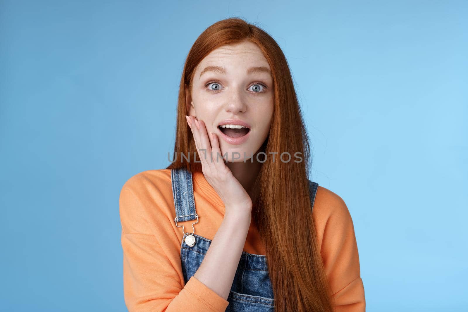 Amazed friendly sensitive impressed redhead girl learn awesome pleasant news open mouth wondered astonished smiling glad friend touch cheek amused, standing blue background happy. Copy space