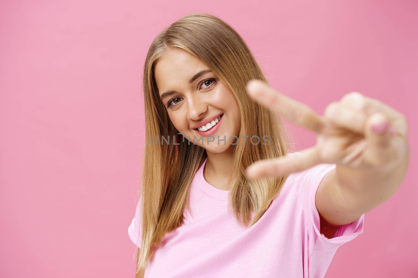 Peaceful and joyful good-looking young girl with straight blond hair in t-shirt pulling hand with peace sign towards camera, tilting head smiling friendly posing against pink background.