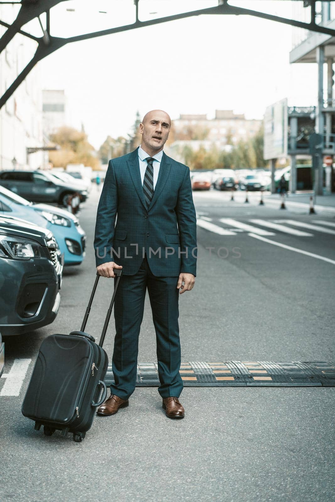 Waiting for his driver or cab handsome businessman stands on parking lot carrying suitcase. Businessman in suit and suitcase on business trip. Man standing outdoors with luggage.