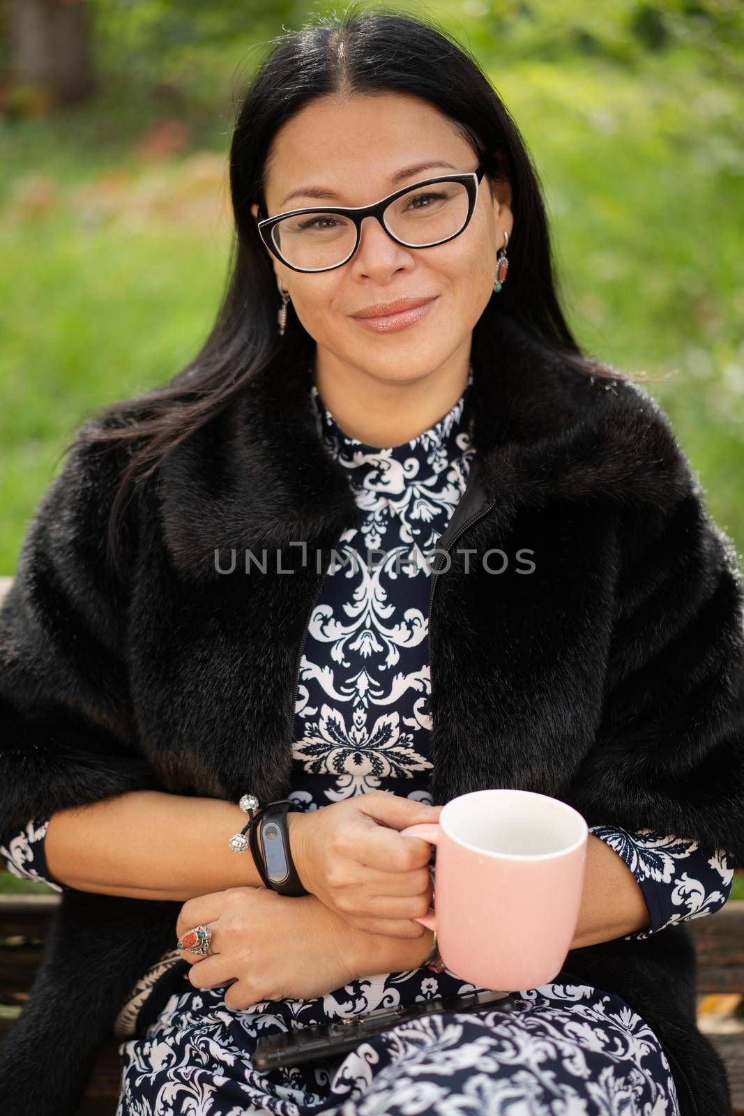 Close up charming asian woman in fur coat and beautiful dress sits at park on old rusty bench with a pinkish cup of coffee or tea. Resting woman in sunny day outdoors by LipikStockMedia