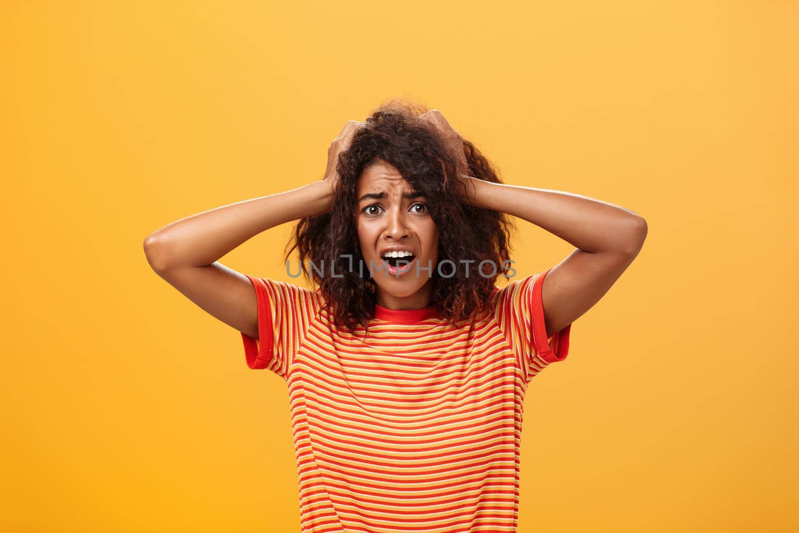 Waist-up shot of concerned shocked and troubled african-american woman with afro hairstyle holding hands on hair frowning looking worried and itnense at camera feeling stressed and nervous. Copy space