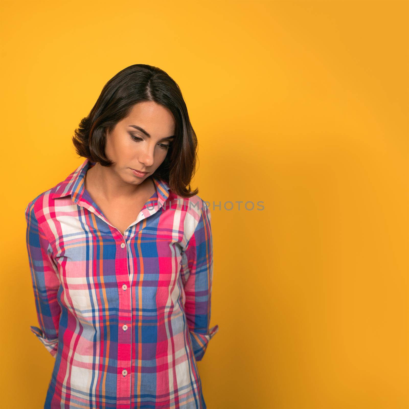 Young Sad pensive woman feels guilty, thinks looking down while standing against of yellow wall in background with copy space on right side by LipikStockMedia