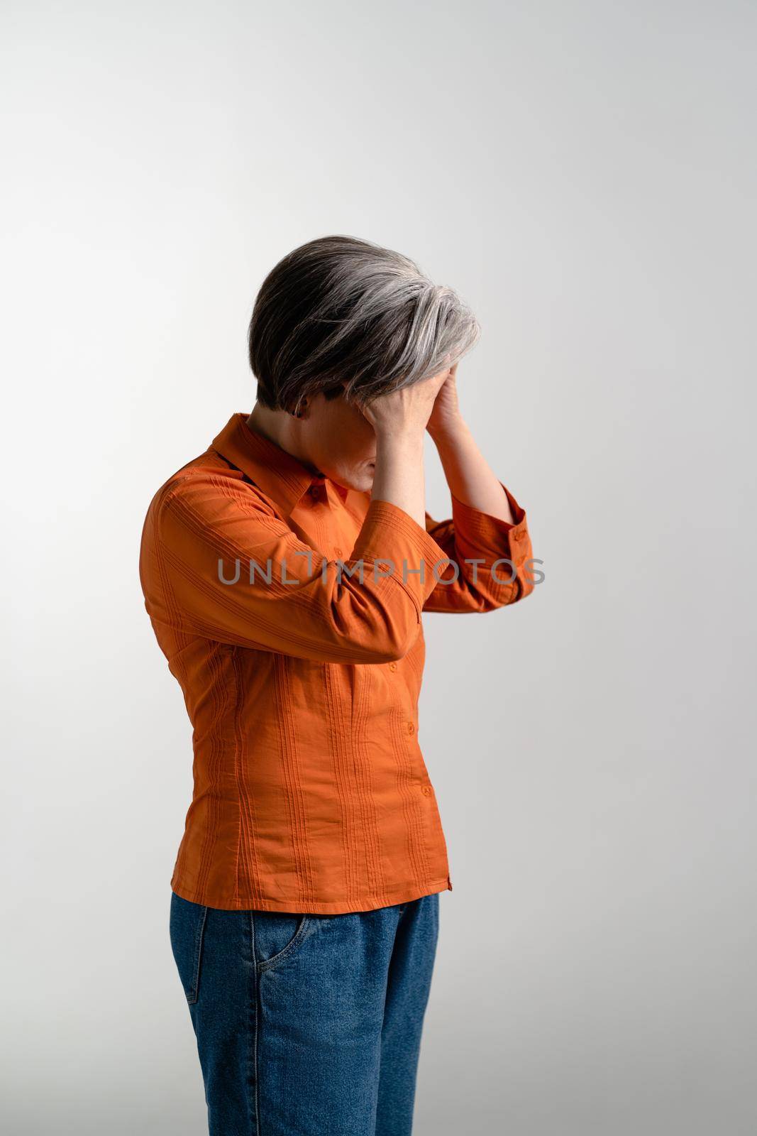 Dejected or depressed mature grey haired woman in orange shirt looks down holding her forehead with two hands. Pretty mid aged grey haired woman in orange shirt isolated on grey background by LipikStockMedia