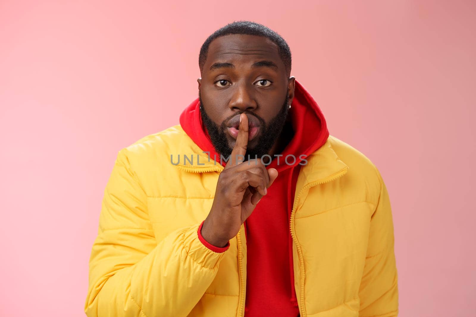 Waist-up serious-looking african-american bearded man in yellow jacket red hoodie bend camera asking keep quiet say shhh show shush gesture index finger on lips, confidential information taboo by Benzoix