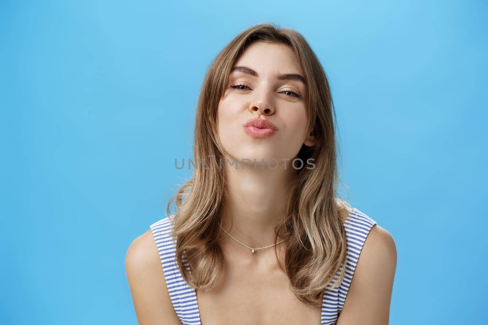 Close-up shot of attractive sensual and flirty young cute woman folding lips in mwah bending towards camera smiling and giving kiss at camera standing happy and self-assured against blue background. Body language concept
