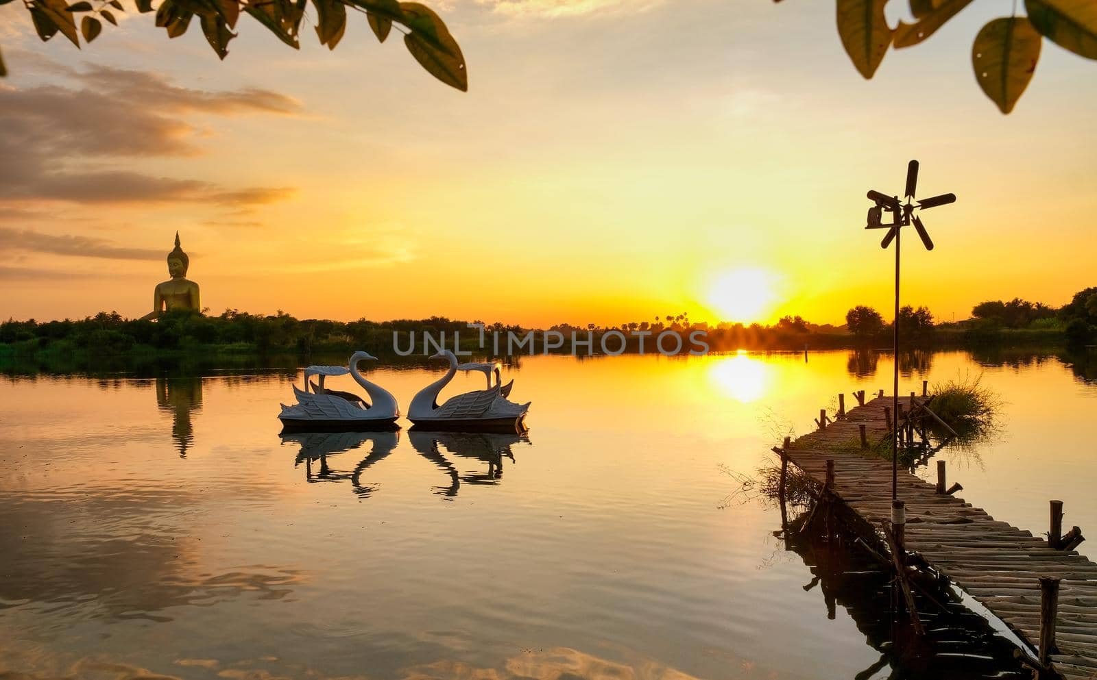 Landscape of lake with wood bridge and reflection of big buddha, goose boat and sunset during evening with yellow and orange light.