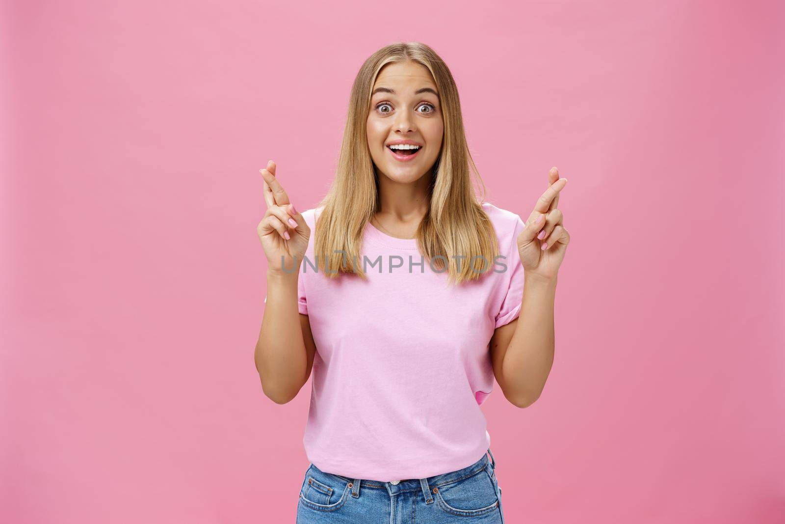 Amused hopeful and delighted attractive female student with tanned skin and blond hair in t-shirt gasping amused and thrilled crossing fingers for good luck wishing and cheering for wish come true. Body language concept