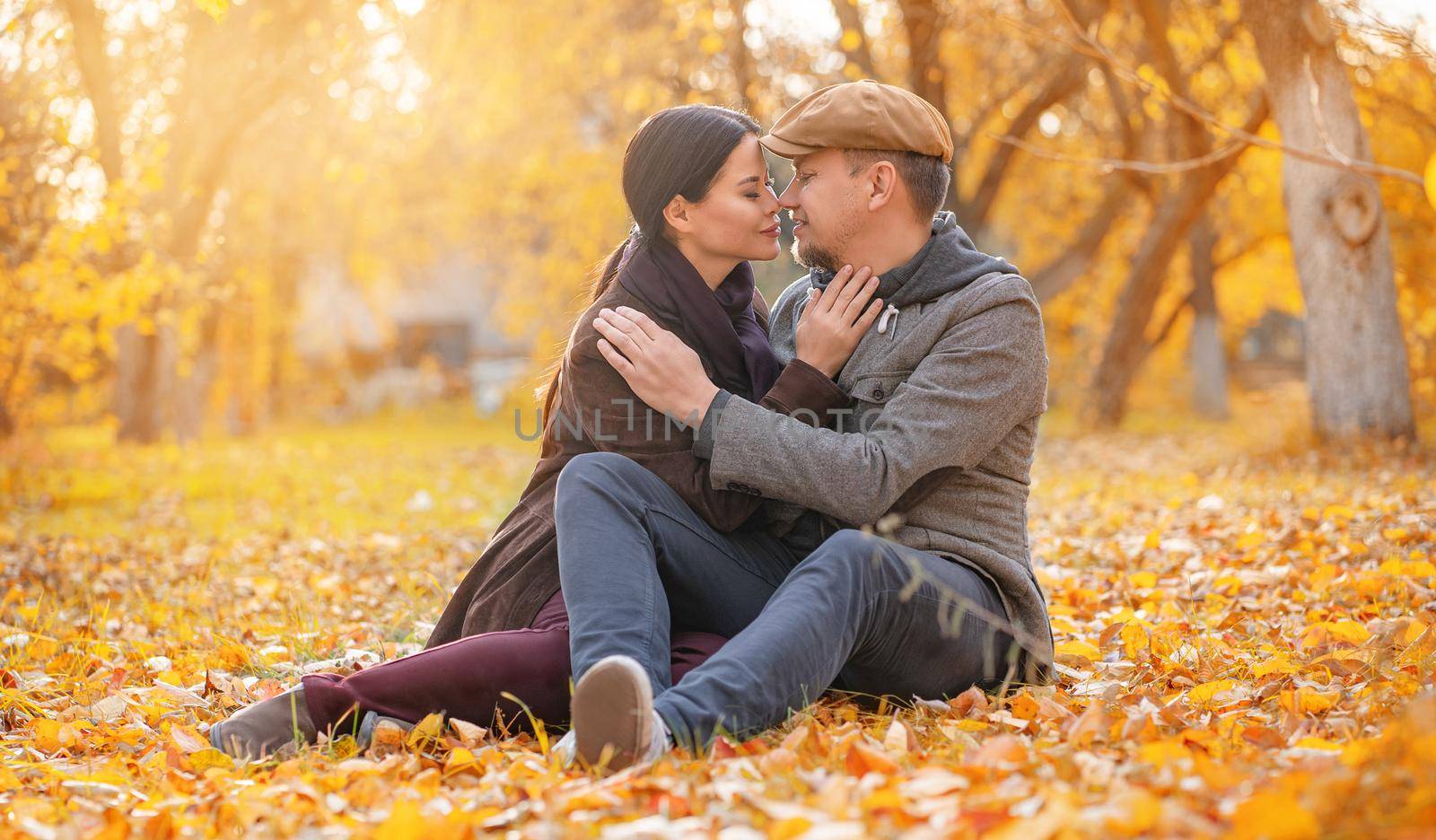 Romantic Couple Sitting on Fall Foliage Nose To Nose With Closed Eyes in Autumn Suburb. Nice Man Hugs His Beloved Woman. Casual Style. Close-up. Fall Background. High quality photo