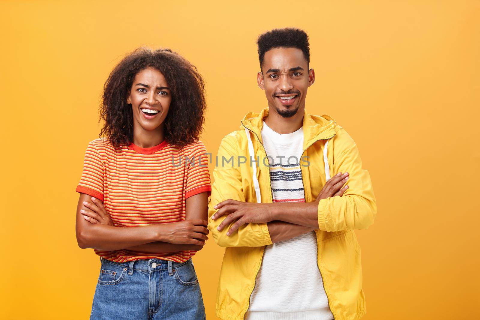 Portrait of surprised sarcastic african american woman with afro hairstyle standing with cute boyfriend crossing arms on chest laughing from scorn and fun over orange background by Benzoix