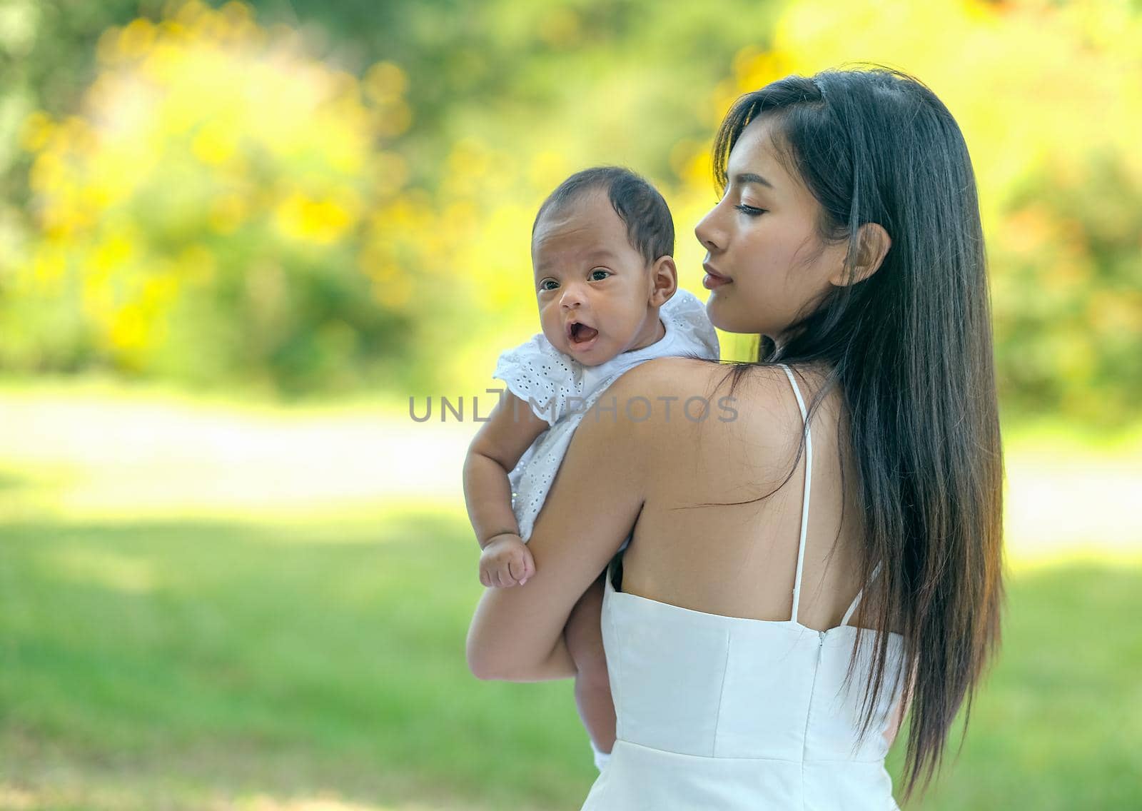 Young Asian beautiful mother carry her little baby and the baby look forward in the green garden with morning light.
