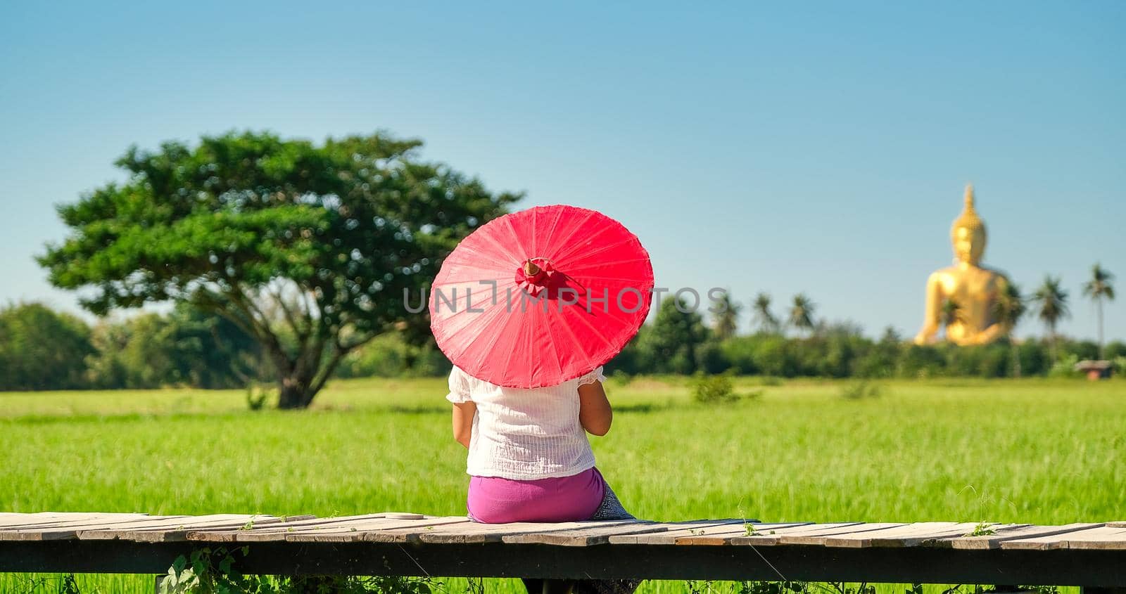 Woman with traditional skirt and red umbrella sit on wood bridge in front of green rice field and she look to Buddha statue with day light and blue sky.