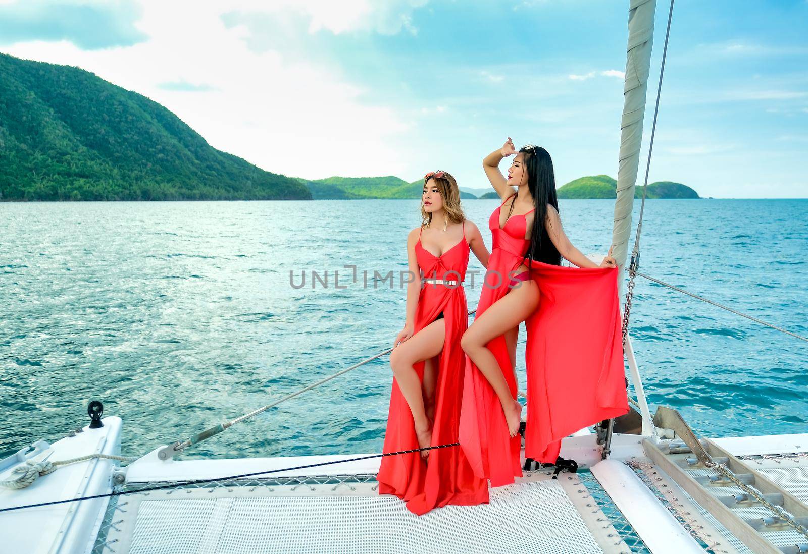 Two of Pretty Asian women with red dress stand on bow of yacht during boat trip during day time.