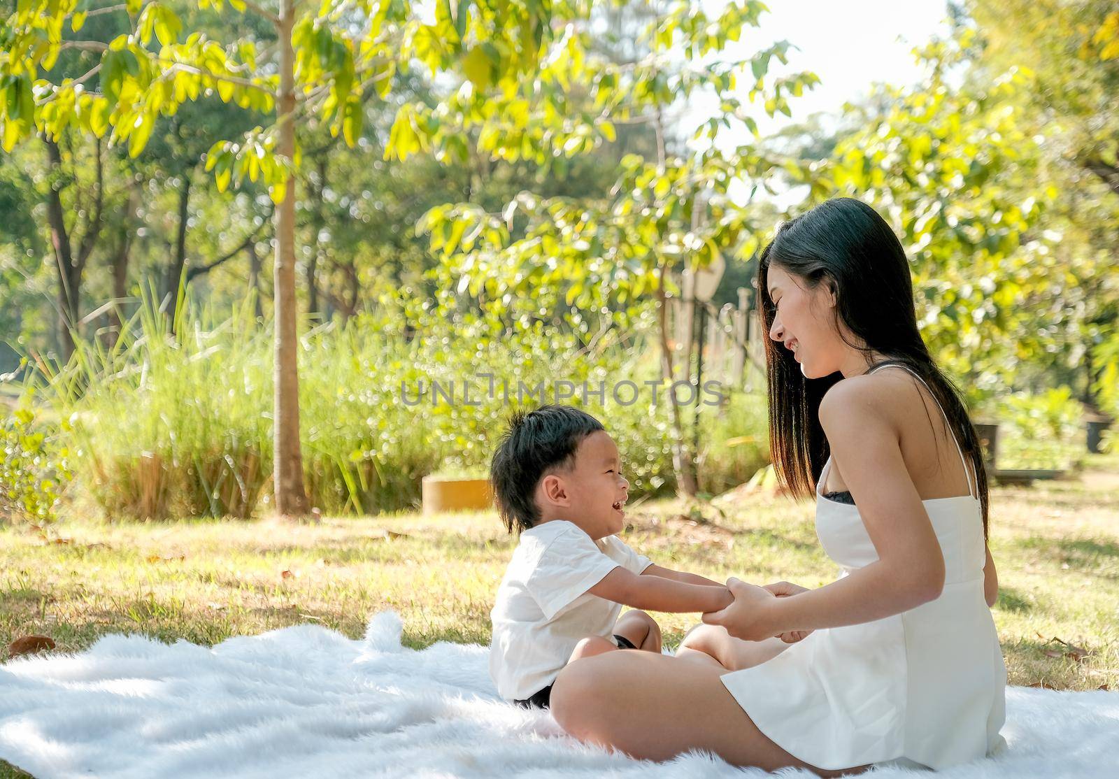 Young Asian mother play and fun with her little boy on white carpet in the green garden with morning light.