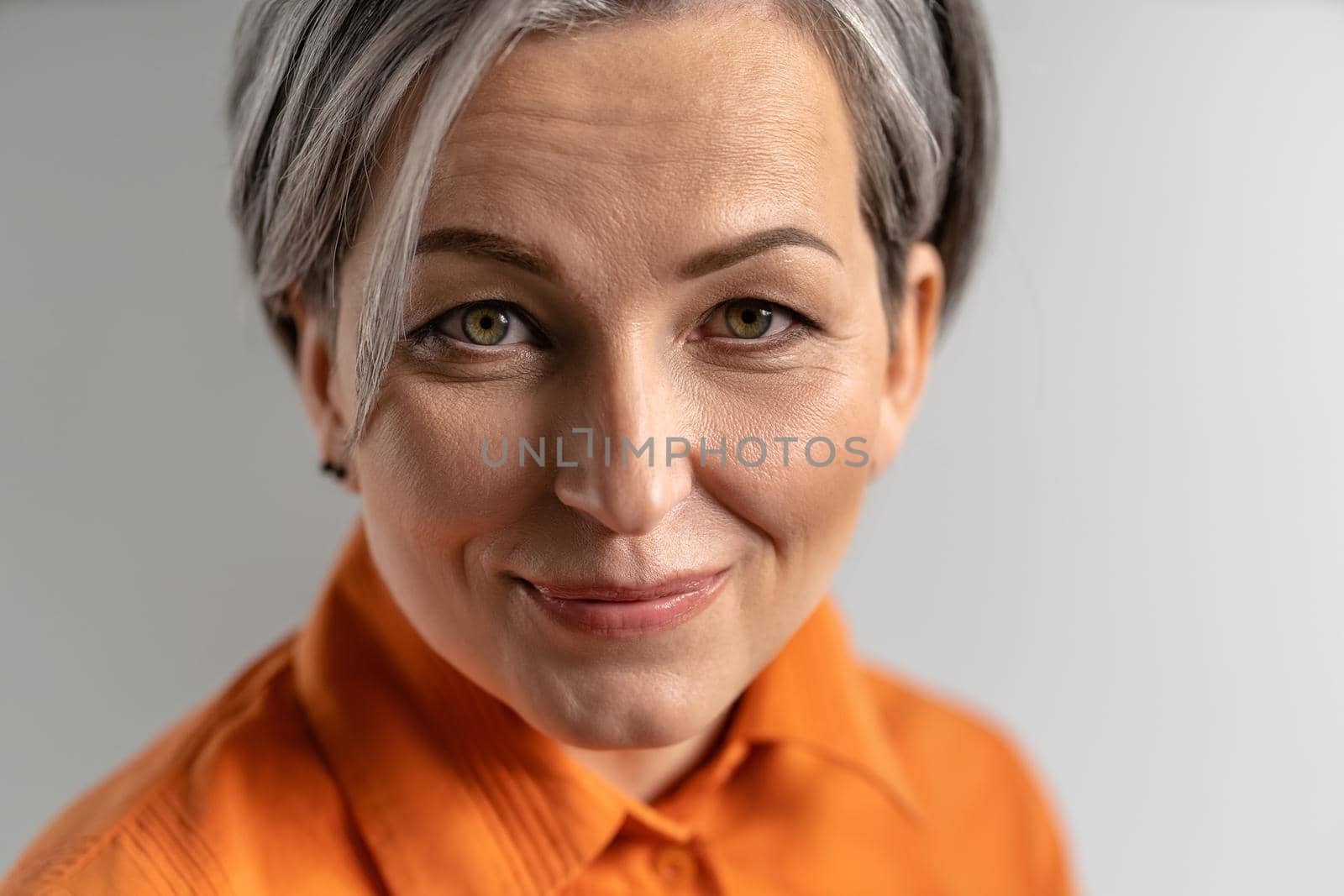 Close up portrait of a happy mature grey haired woman wearing orange shirt looking at camera. Pretty mid aged grey haired woman in orange shirt isolated on grey background.