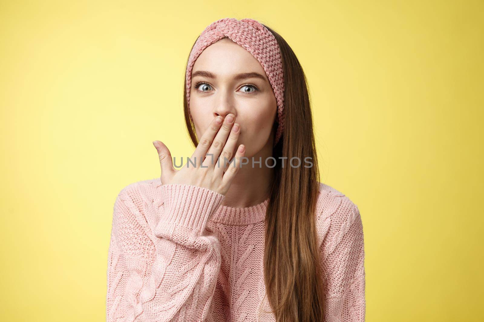 Talkative young cute office worker wearing sweater, knitted headband holding palm on mouth surprised and amazed, learning interesting shocking rumor, gossiping entertained over yellow wall.