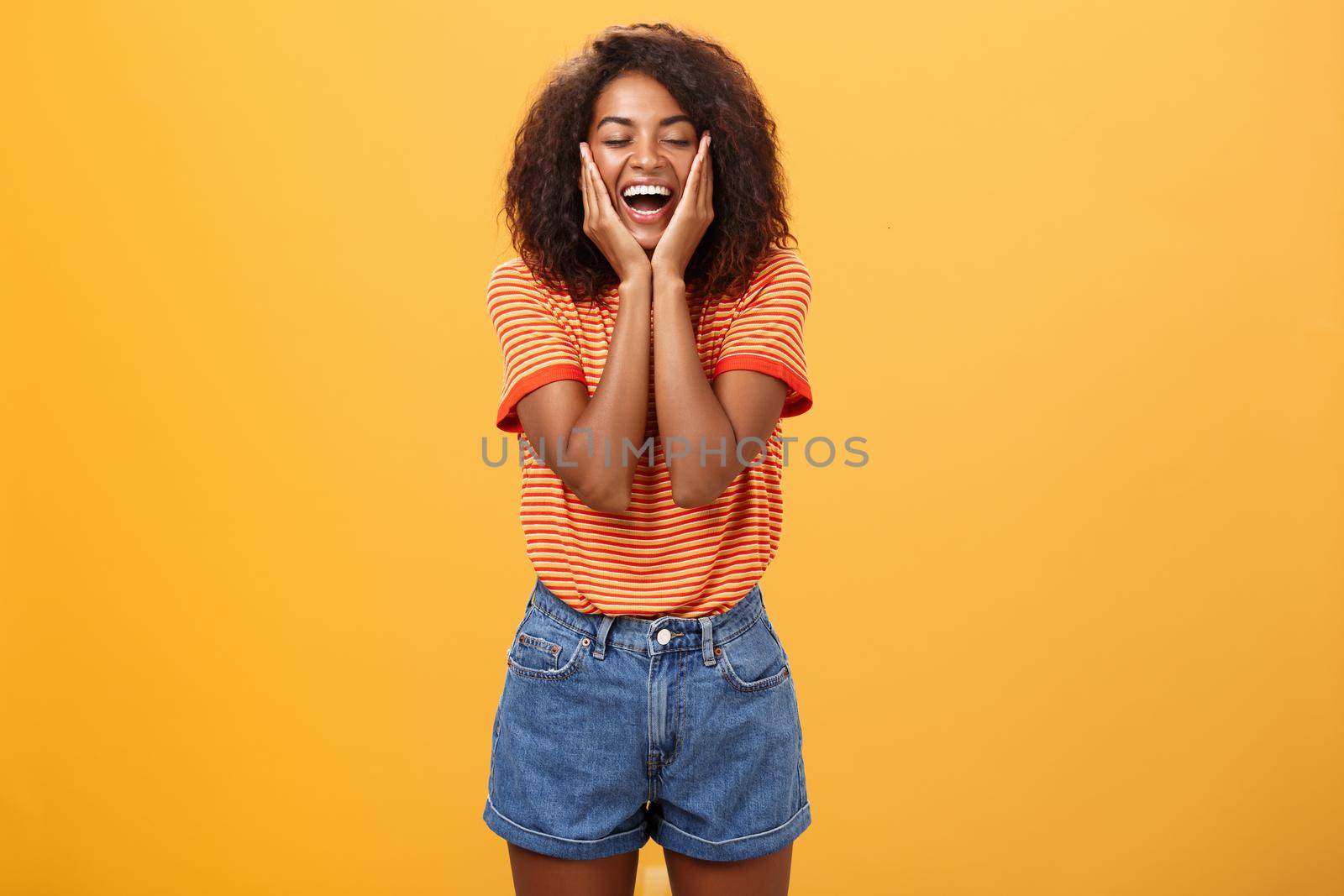 Woman happy and delighted with great result after curing acne with skincare products touching cheeks smiling happily with closed eyes feeling pleased and amused over orange background. Copy space