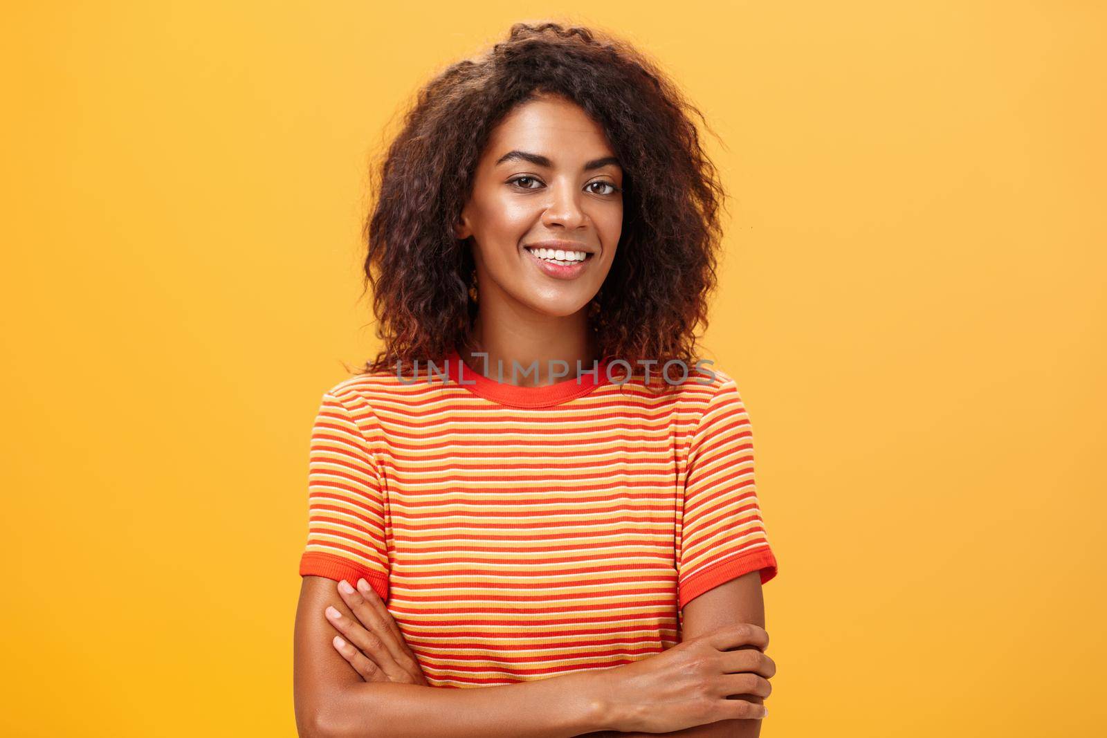 Waist-up shot of creative ambitious good-looking female coworker in trendy striped t-shirt holding hands crossed on chest smiling broadly at camera friendly and self-assured over orange background. Lifestyle.