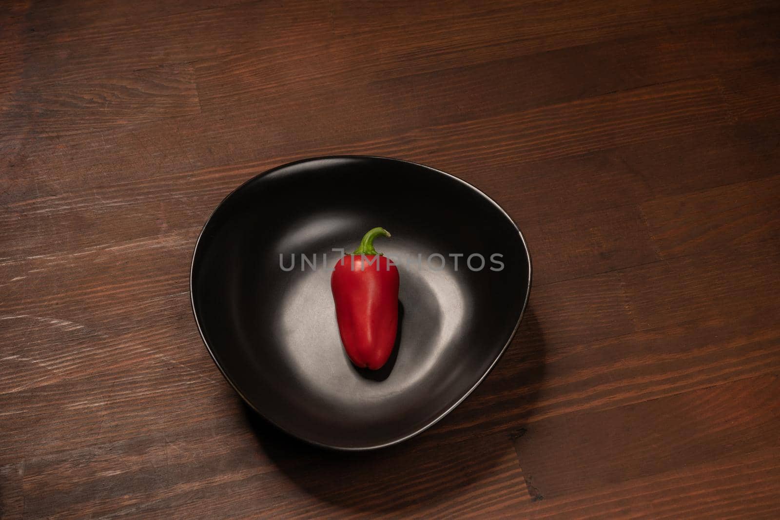 Organic mini red bell pepper laying lonely in a deep black bawl and ready to eat isolated on wooden background.