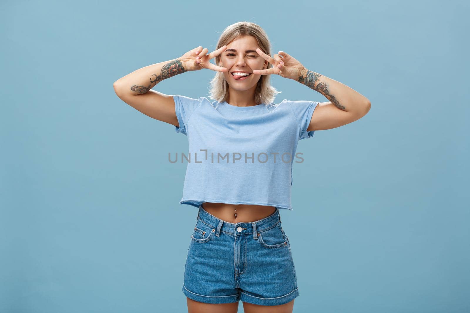 Enjoy life and dream. Portrait of joyful good-looking carefree european female student with blond short haircut winking sticking out tongue entertained and showing peace signs over eyes near blue wall by Benzoix